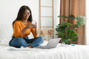 African American Woman Using Phone And Laptop Sitting In Bedroom