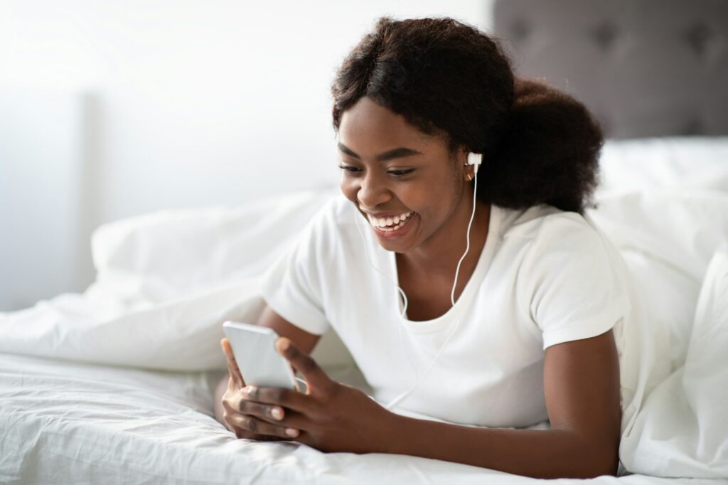 Happy young black woman laying in bed with smartphone