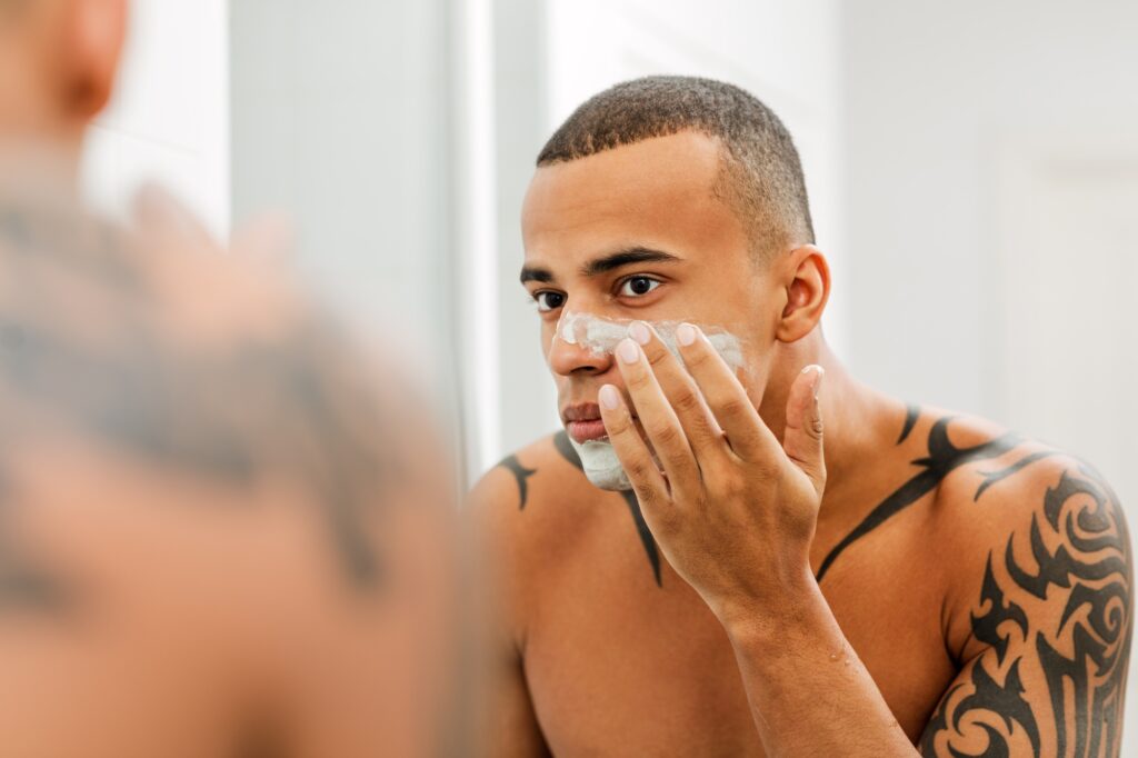 African man in bathroom looking into the mirror and applying natural mask on his face