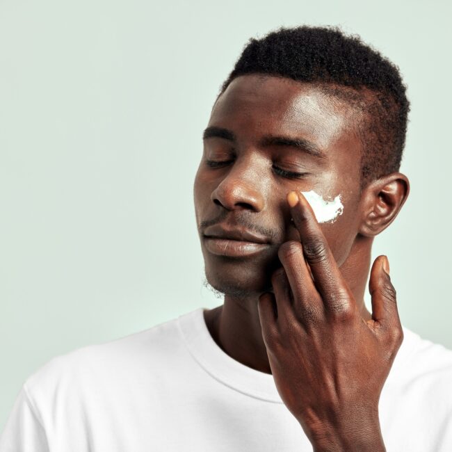 Black guy with youthful skin applying a moisturizing cream on his face to keep his skin soft and