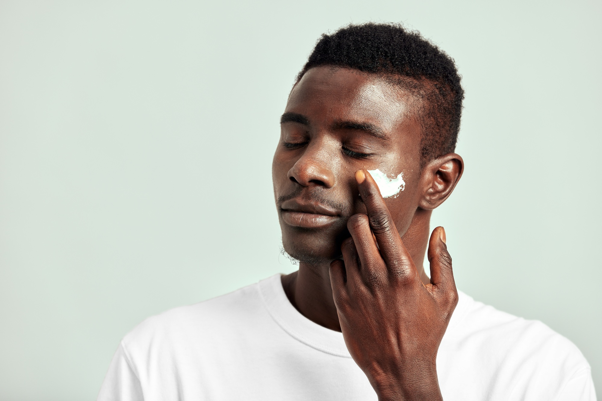 Black guy with youthful skin applying a moisturizing cream on his face to keep his skin soft and