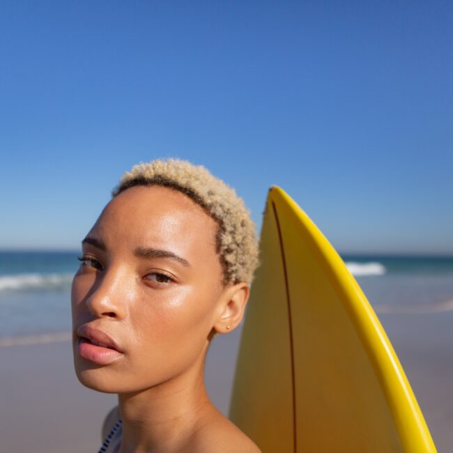 Close-up of beautiful African american woman standing with surfboard on beach in the sunshine