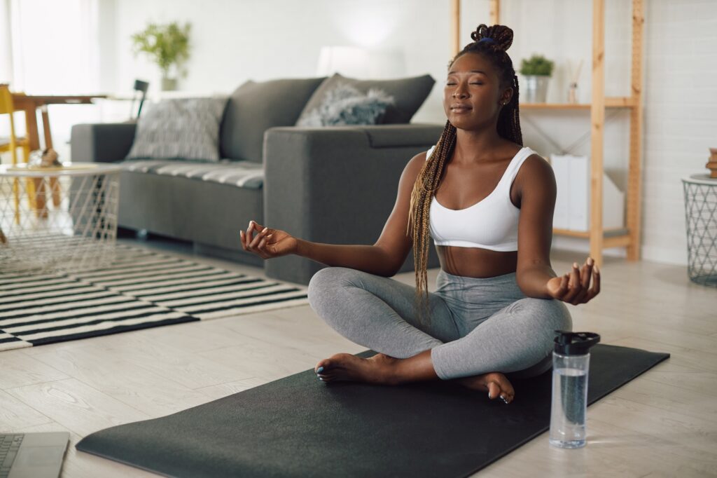 Young African American woman meditating in lotus pose while working out at home.